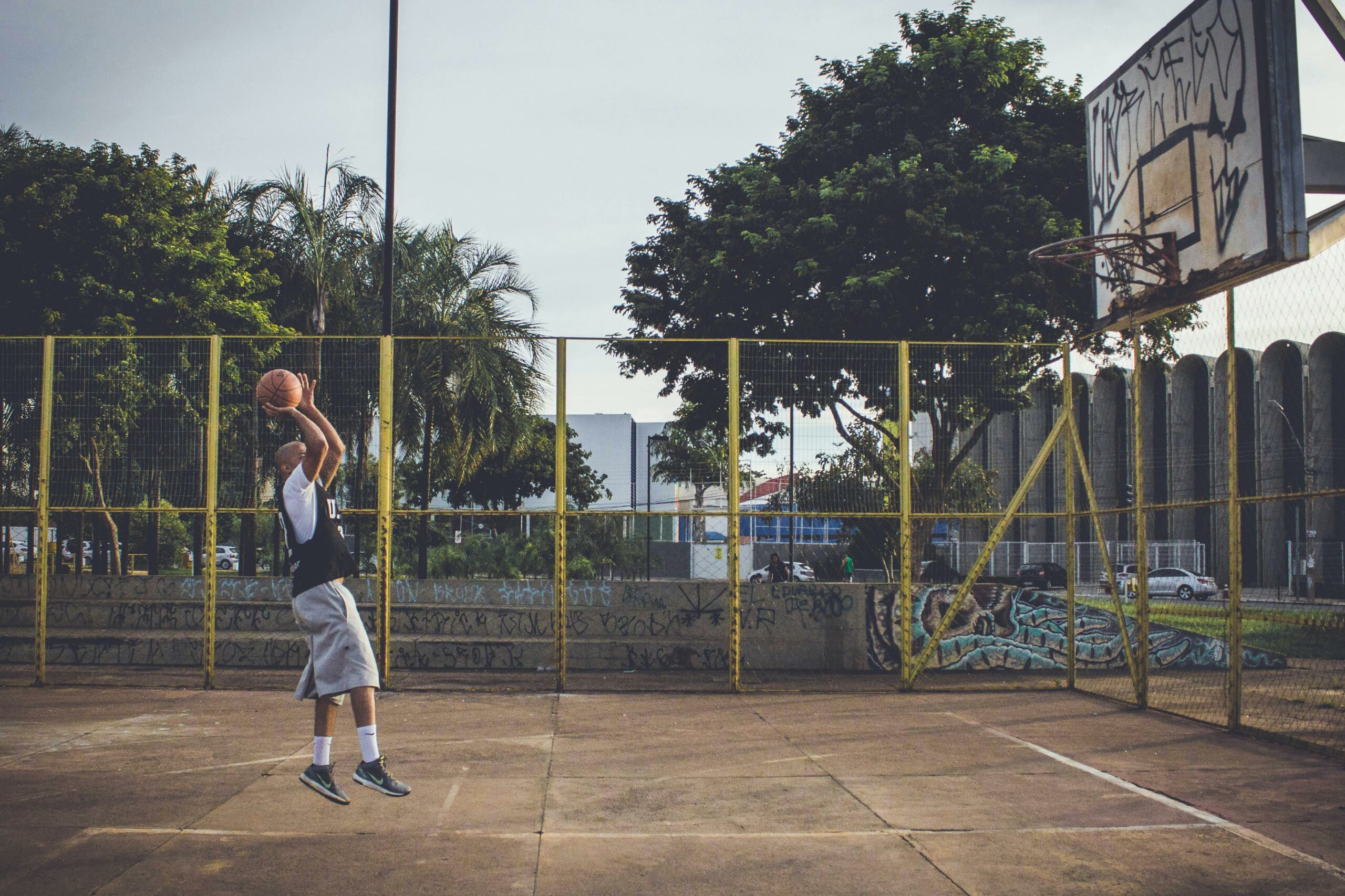 A man in athletic wear leaps for a basketball shot on an outdoor urban court.